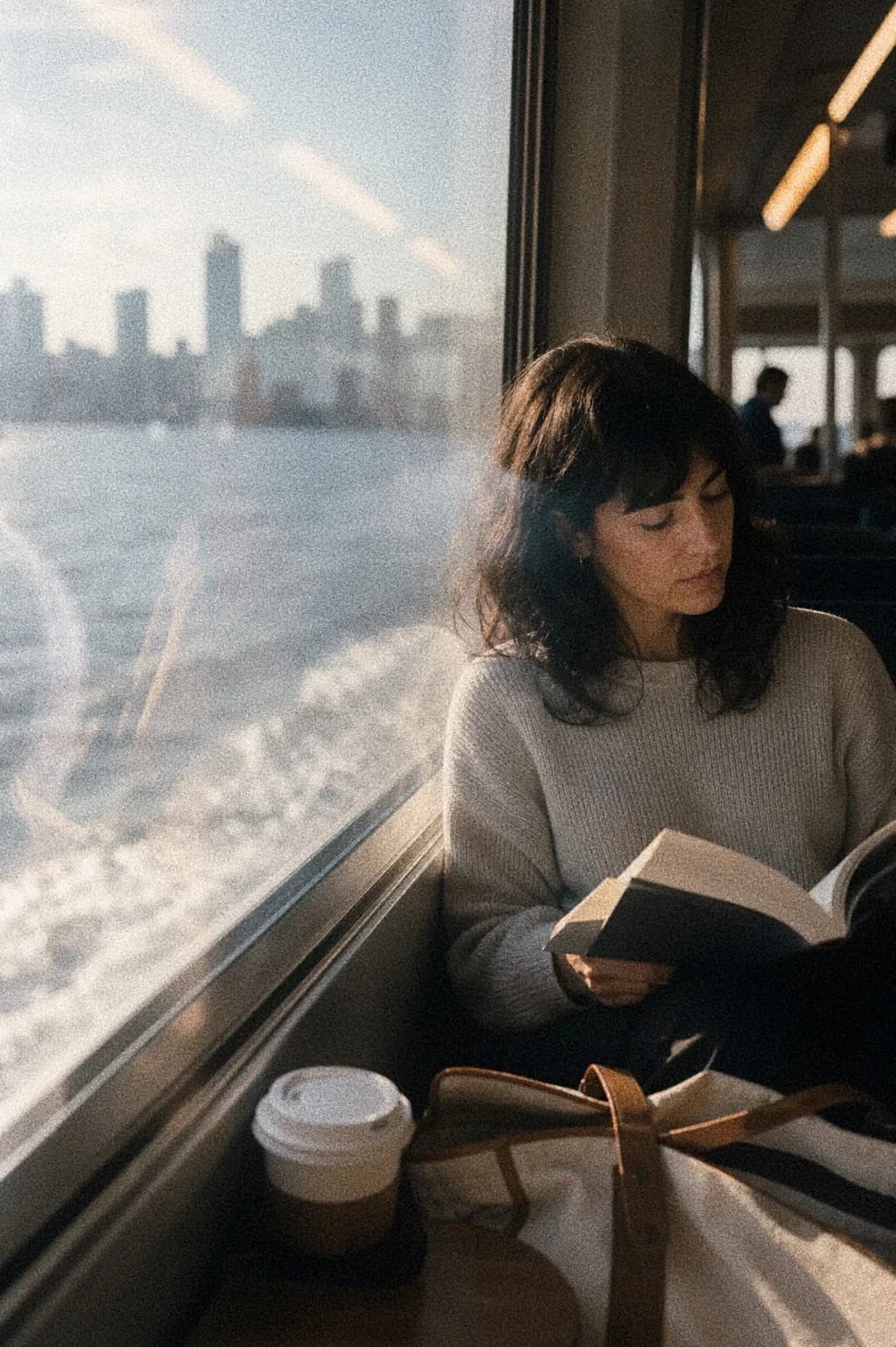 Person reading on a ferry with city water outside the window.